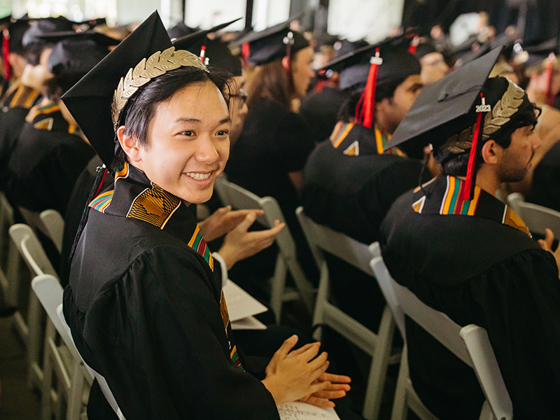 A graduate in a cap and gown turns and smiles while sitting next to other graduates during Commencement.