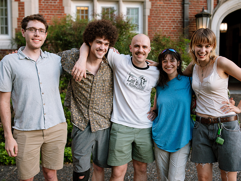 Five alumni pose for a photo while smiling with their arms wrapped around each other.