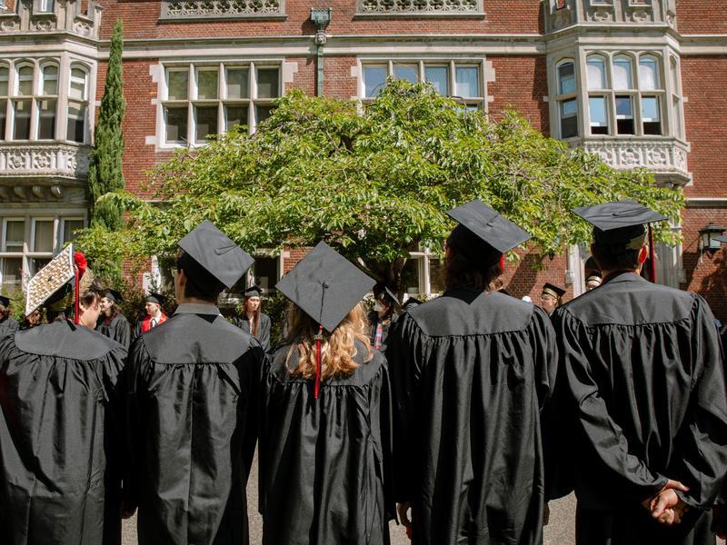 Five graduates in caps and gowns stand shoulder-to-shoulder with their backs facing the camera and look up at Old Dorm Block.